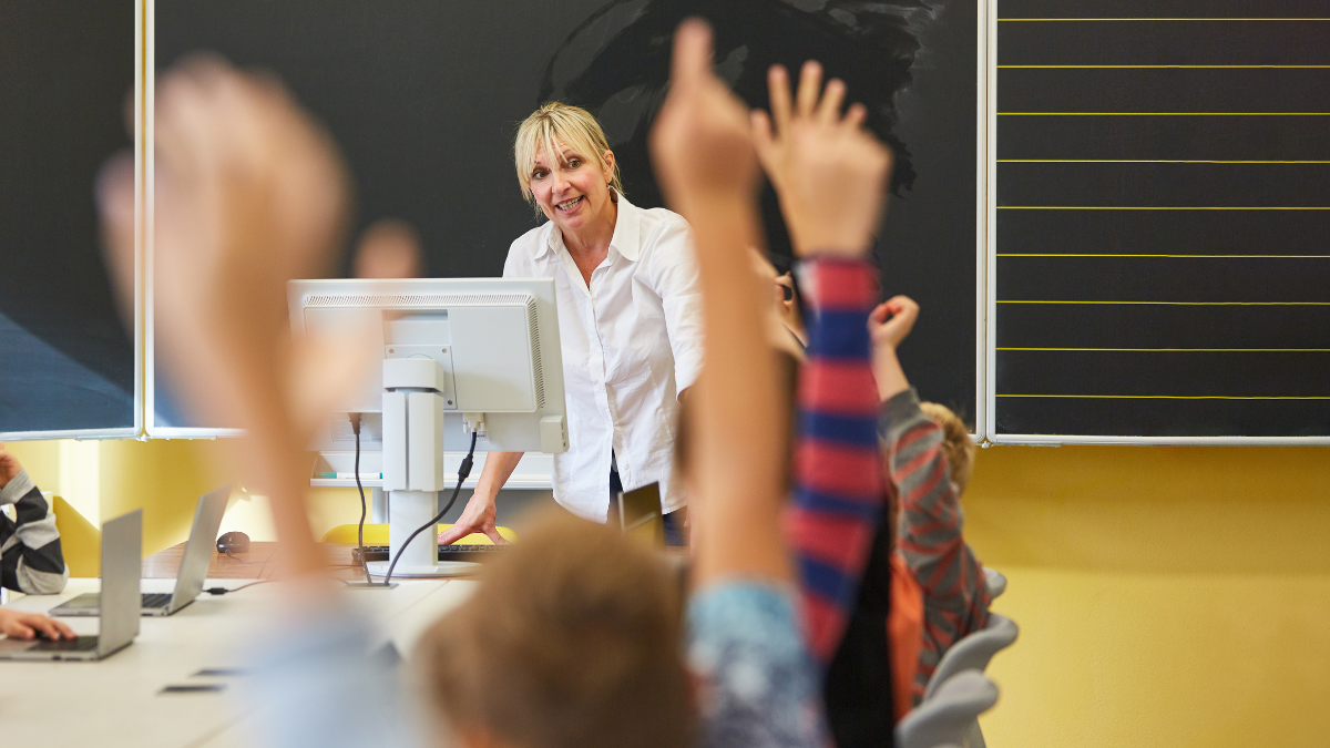 Uma sala de aula com uma Pedagoga e alunos ao redor de uma mesa com notebbok - Fonte Canva